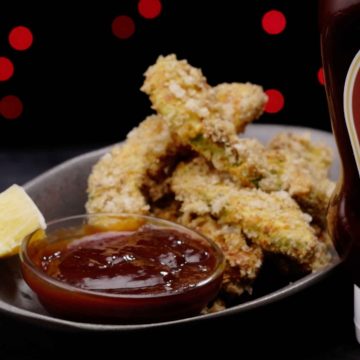 Golden breaded avocado fries on a dark plate with barbecue dipping sauce, red bokeh lights in background
