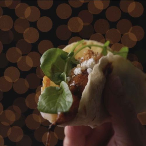Hand holding a soft taco filled with glazed carrots, greens, and crumbled cheese against a bokeh background