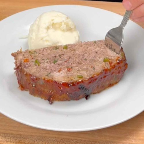 Slice of spicy meatloaf with visible peppers and vegetables on white plate with mashed potatoes and fork