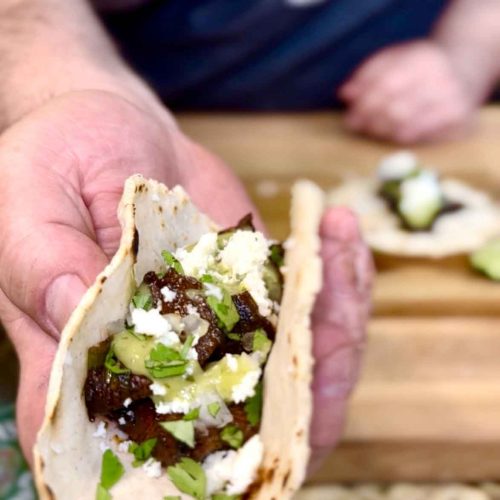 Person holding a carne asada taco with dark meat, white cheese, green herbs, and salsa in a soft tortilla
