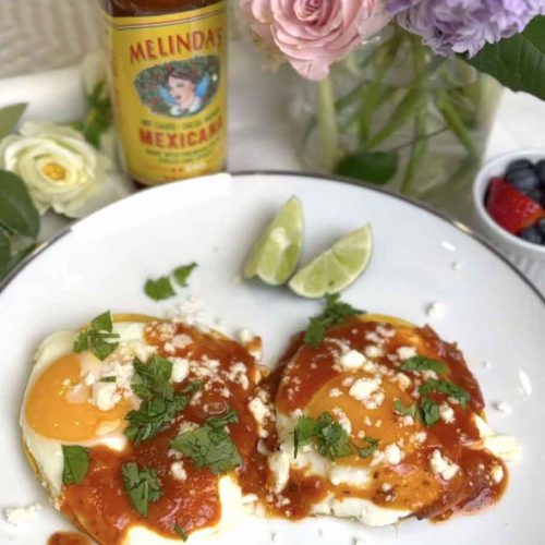 Two fried eggs topped with red salsa, white cheese crumbles, and cilantro on a white plate with lime wedges