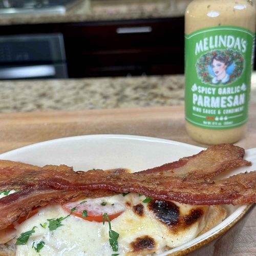 Crispy bacon strips on melted cheese dish with Melinda's Spicy Garlic Parmesan sauce bottle in background