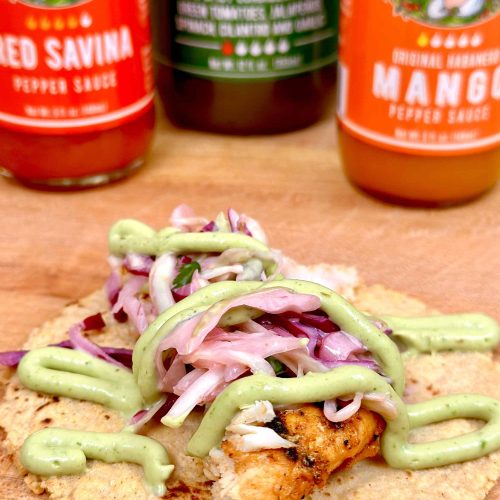 Fish taco with grilled fish, purple cabbage slaw, and green avocado crema drizzle, with hot sauce bottles in background