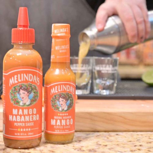 Two bottles of Melinda's Mango Habanero hot sauce on a counter with someone preparing a cocktail in the background.