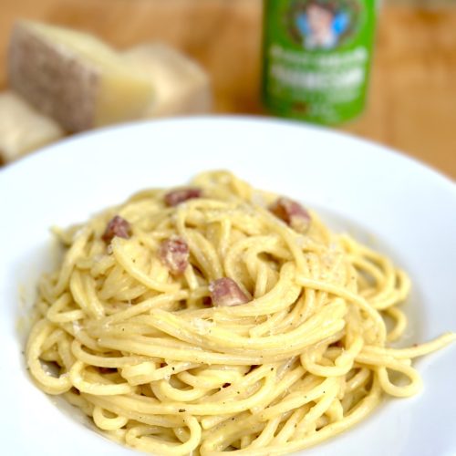 Plate of spaghetti carbonara with bacon pieces, creamy sauce, and black pepper on white dish with cheese and beer bottle