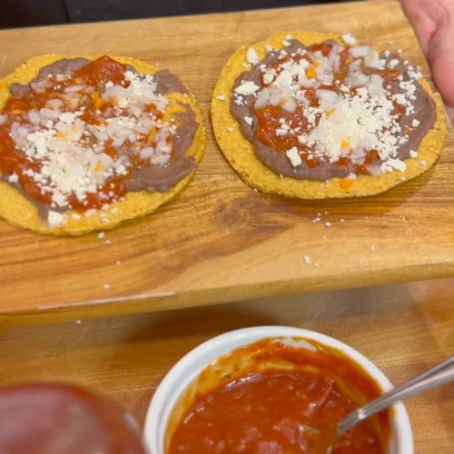 Two golden corn tortillas topped with refried beans, red sauce, and crumbled cheese on a wooden board with sauce bowl