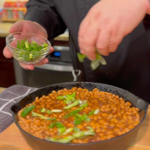 Chef adding fresh green onions to a cast iron skillet filled with bacon chipotle baked beans