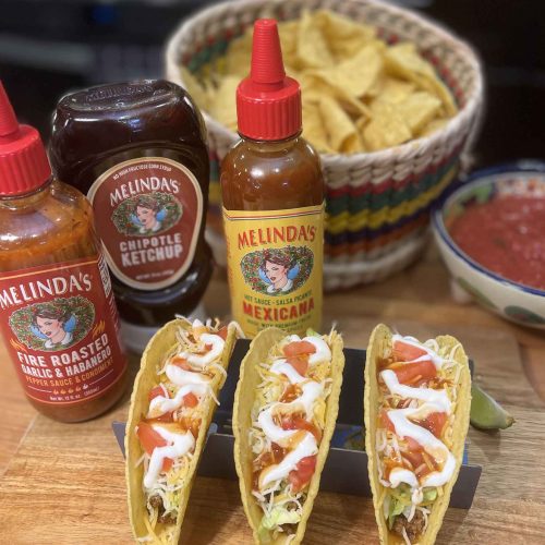 Three hard shell tacos with toppings alongside Melinda's hot sauce bottles, tortilla chips, and salsa on wooden surface.