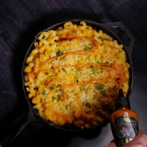 Golden mac and cheese baked in cast iron skillet with crispy top and herbs, bottle of Melinda's hot sauce beside it