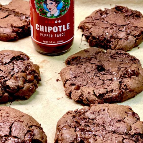 Dark chocolate cookies with cracked surfaces arranged on parchment paper next to a bottle of Chipotle pepper sauce