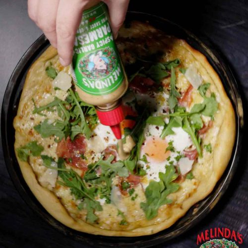 Hand drizzling Melinda's hot sauce over breakfast pizza topped with eggs, arugula, and cheese in cast iron skillet