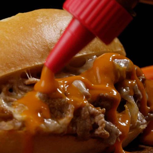 Philly cheese steak sandwich with melted cheese and sautéed onions, hot sauce being drizzled on top