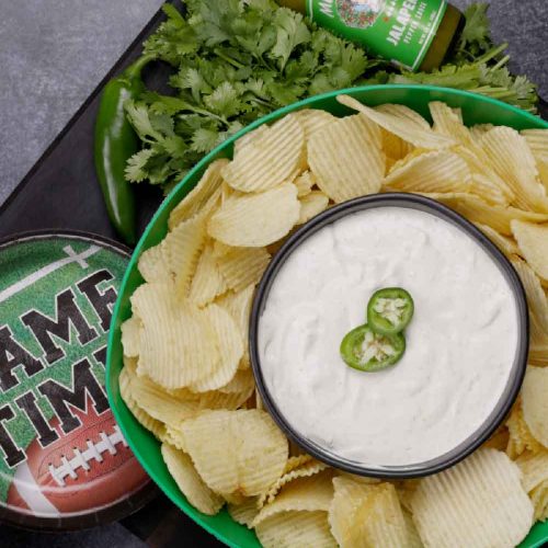 Bowl of jalapeño sour cream dip garnished with jalapeño slices, surrounded by potato chips and cilantro on game day plate