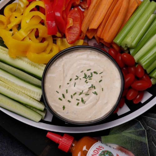 Vegetable platter with colorful bell peppers, carrots, celery, and cherry tomatoes surrounding a bowl of creamy buffalo...