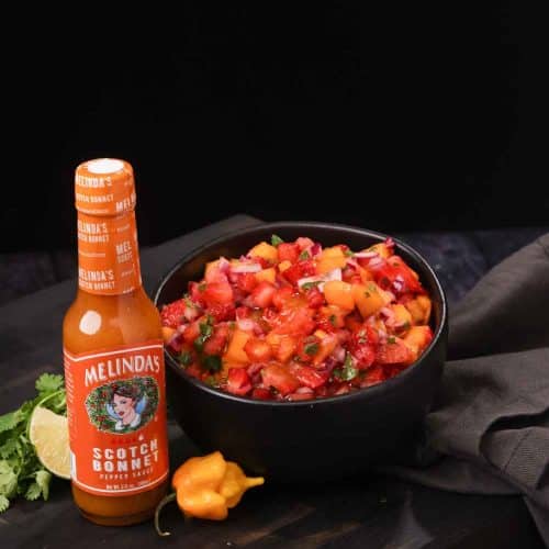 Bowl of colorful fruit salsa with diced peppers and herbs next to Melinda's Scotch Bonnet hot sauce bottle on dark background