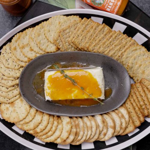 Cream cheese topped with mango honey glaze on dark plate, surrounded by round crackers with hot sauce bottle visible
