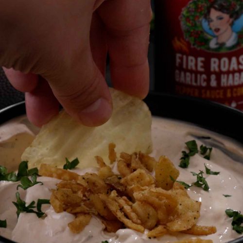 Hand dipping chip into creamy white dip garnished with crispy fried onions and herbs on white plate
