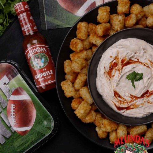 Golden tater tots arranged around a bowl of ranch dip with hot sauce swirl, alongside Melinda's Chipotle sauce bottle