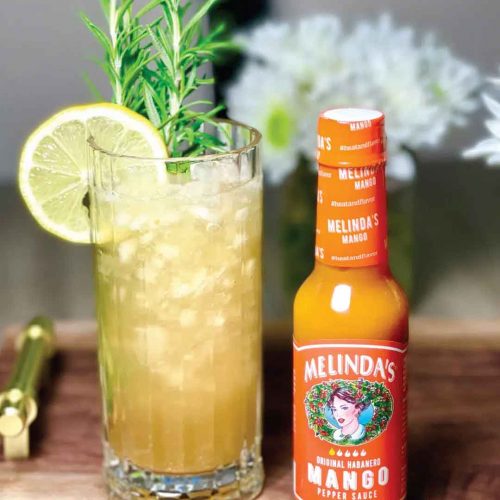 Tall glass of spicy mango rosemary lemonade with lime wedge and rosemary garnish next to Melinda's Mango hot sauce bottle