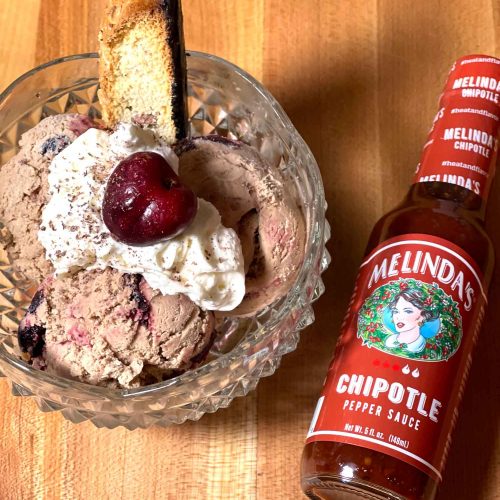 Cherry chocolate chipotle ice cream in glass bowl with whipped cream and cherry, alongside Melinda's Chipotle sauce bottle