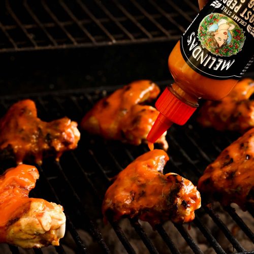 Chicken wings being basted with Melinda's hot sauce on a grill grate, showing the cooking process for spicy wings.