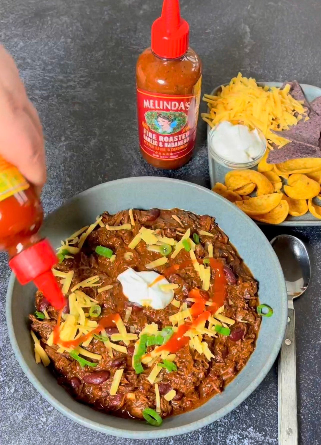 Bowl of chili con carne topped with cheese, sour cream, and green onions, served with tortilla chips and hot sauce