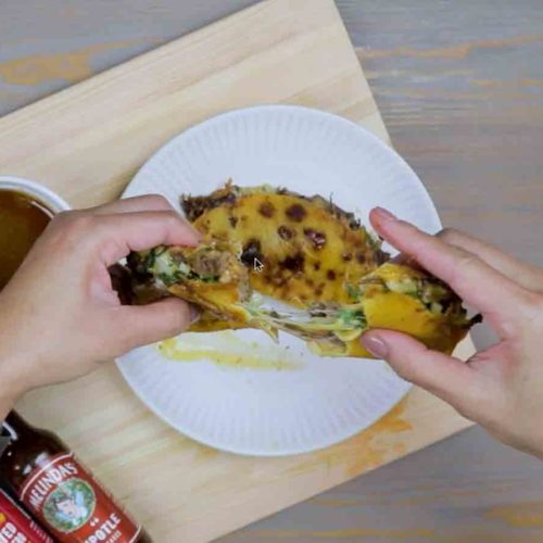 Hands holding a birria taco filled with meat, cheese, and cilantro on a white plate with Melinda's hot sauce bottle visible
