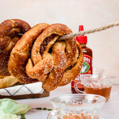 Large twisted pretzel with coarse salt alongside hot sauce bottle and honey mustard dipping sauce