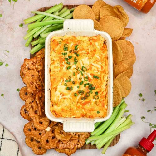 Buffalo chicken dip in white baking dish surrounded by celery sticks, pretzel crisps, and tortilla chips