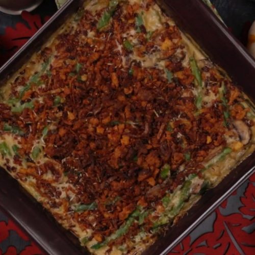 Square baking dish filled with green bean casserole topped with crispy fried onions and herbs on a red patterned surface.