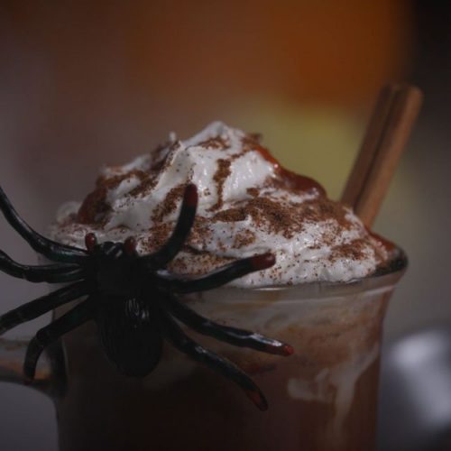 Hot chocolate topped with whipped cream and cocoa powder, garnished with a plastic spider for Halloween decoration