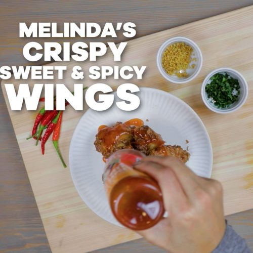 Hand dipping crispy chicken wing in sauce on white plate, with fresh chilies and herb garnishes nearby