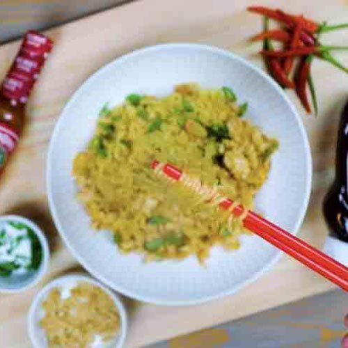Spicy ramen noodles with green herbs in white bowl, red chopsticks, surrounded by hot sauce bottles and chili peppers