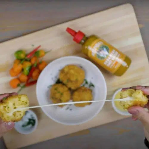 Golden fried mac and cheese balls on white plate with Melinda's hot sauce bottle and colorful vegetables on wooden board