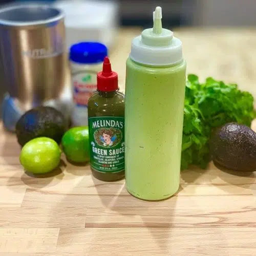 Green squeeze bottle of avocado lime cilantro crema next to Melinda's Green Sauce bottle, limes, cilantro, and avocado