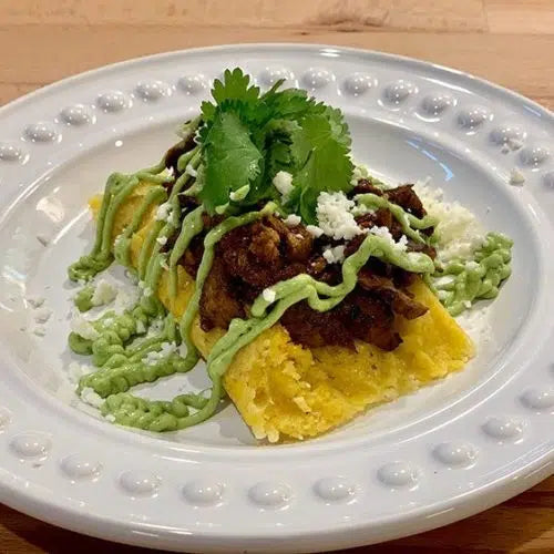 A plated tamalito topped with meat, lettuce, crumbled cheese, and cilantro garnish on a white decorative plate.