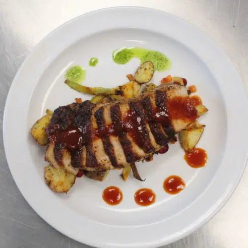 Sliced grilled pork chops with garlic pepper glaze, served with roasted vegetables and green herb sauce on white plate