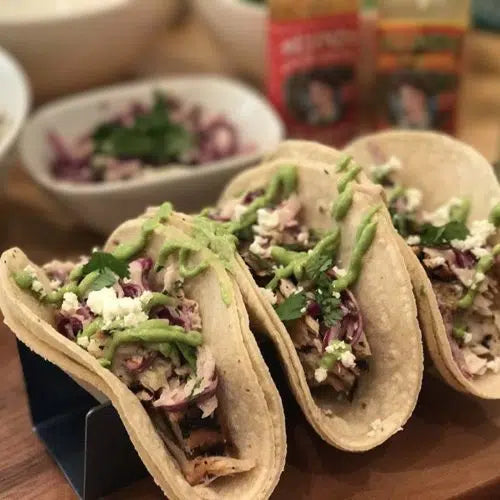 Three fish tacos in corn tortillas topped with purple cabbage, cilantro, cheese crumbles, and green avocado crema