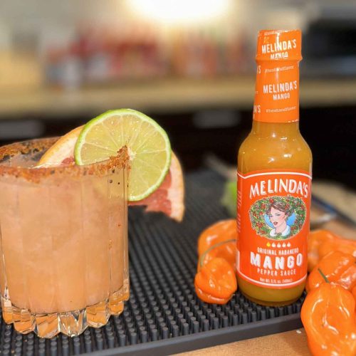 Spicy paloma cocktail with lime garnish next to Melinda's Mango hot sauce bottle and orange habanero peppers on bar mat