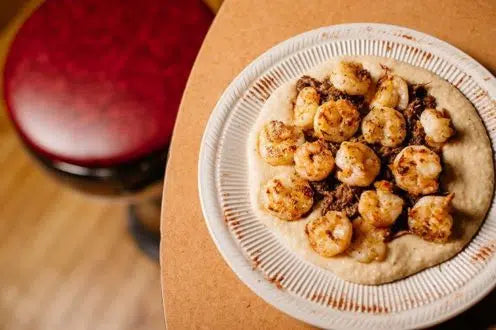 Grilled shrimp served over creamy grits with roasted beef debris on a white ridged plate beside a red wine glass.