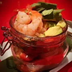 Glass bowl of shrimp cocktail with tropical garnish including lime and herbs on red background