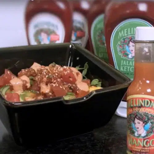 Bowl of poke with diced fish, vegetables, and sesame seeds beside bottles of Melinda's hot sauce on counter