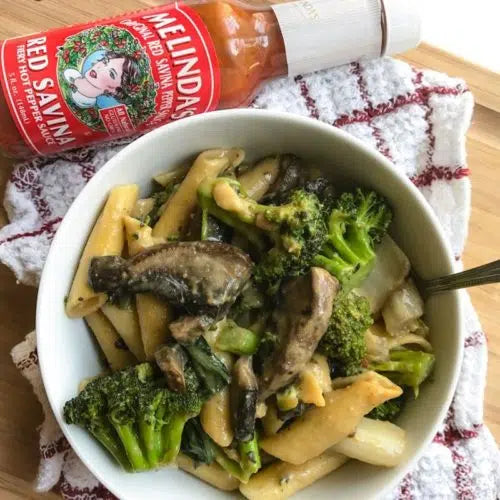 Bowl of pasta with broccoli and mushrooms in peanut sauce, with Melinda's hot sauce bottle on wooden surface