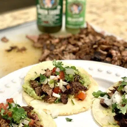 Three corn tortilla tacos filled with seasoned carne asada, diced tomatoes, onions, and fresh cilantro on a white plate