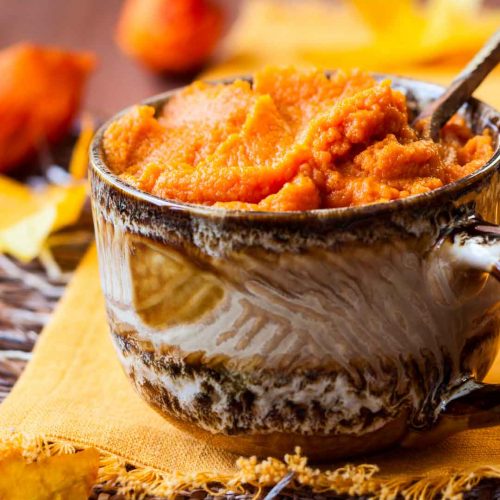 Orange mashed butternut squash in a rustic ceramic mug with wooden spoon, surrounded by fresh oranges on yellow cloth