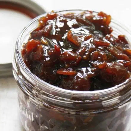 Dark reddish-brown bacon marmalade with visible herbs in an open glass jar with metal lid beside it