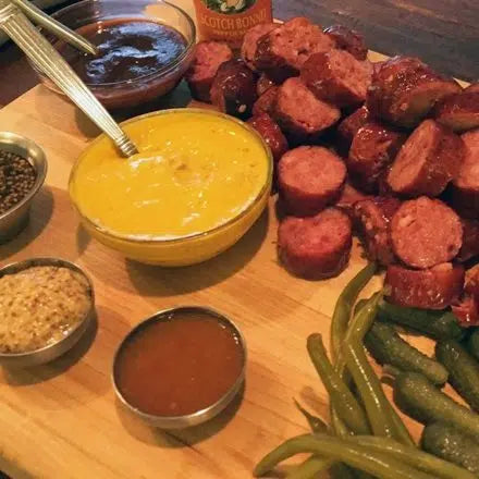 Wooden charcuterie board with sliced sausages, yellow mustard, pickles, and various condiments in small bowls