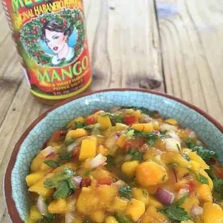 Bowl of colorful mango salsa with diced peppers and herbs, alongside a bottle of Melinda's hot sauce on wooden surface