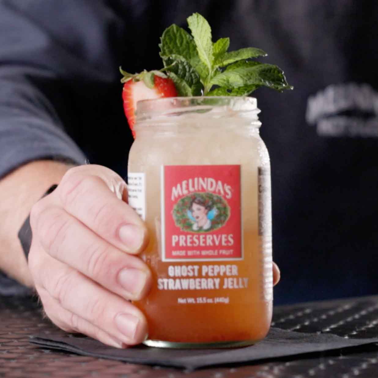 Hand holding mason jar cocktail with Melinda's Ghost Pepper Strawberry Jelly label, garnished with mint and strawberry slice