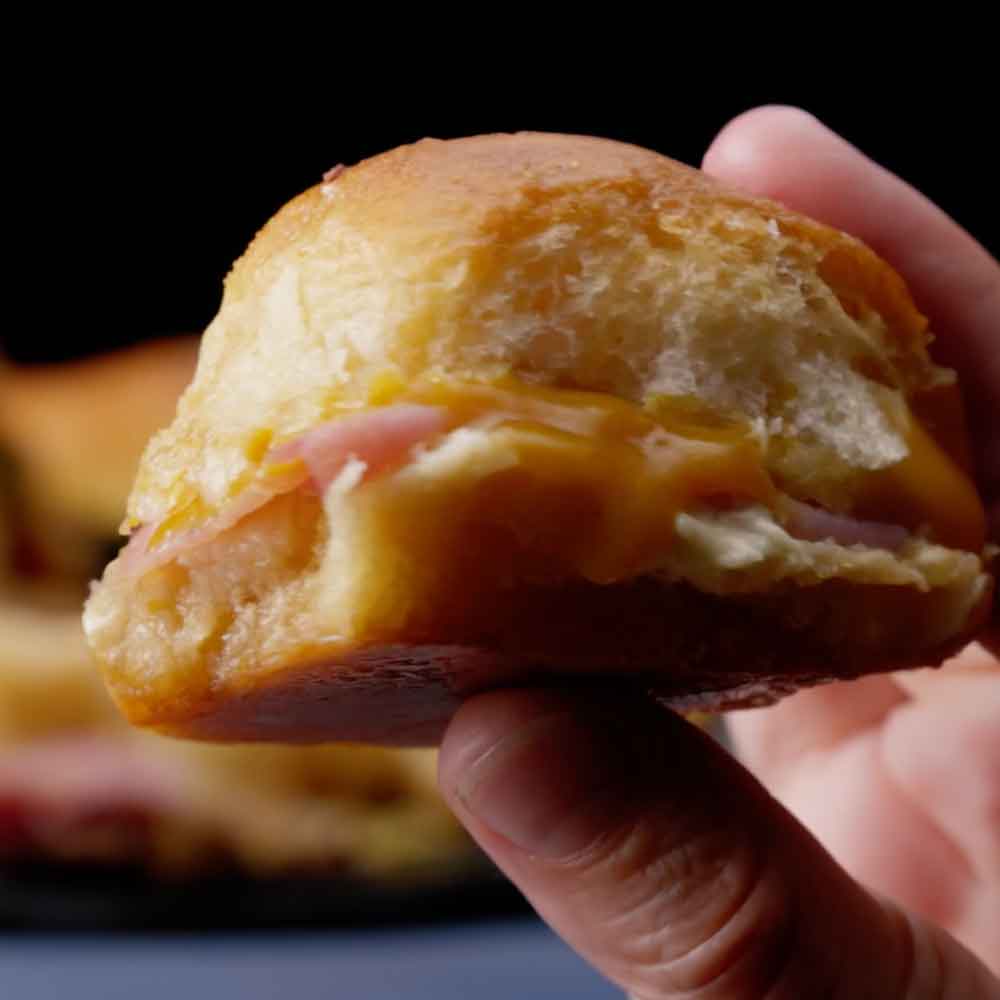 Hand holding a golden dinner roll filled with ham and melted Swiss cheese, pulled apart to show the layers
